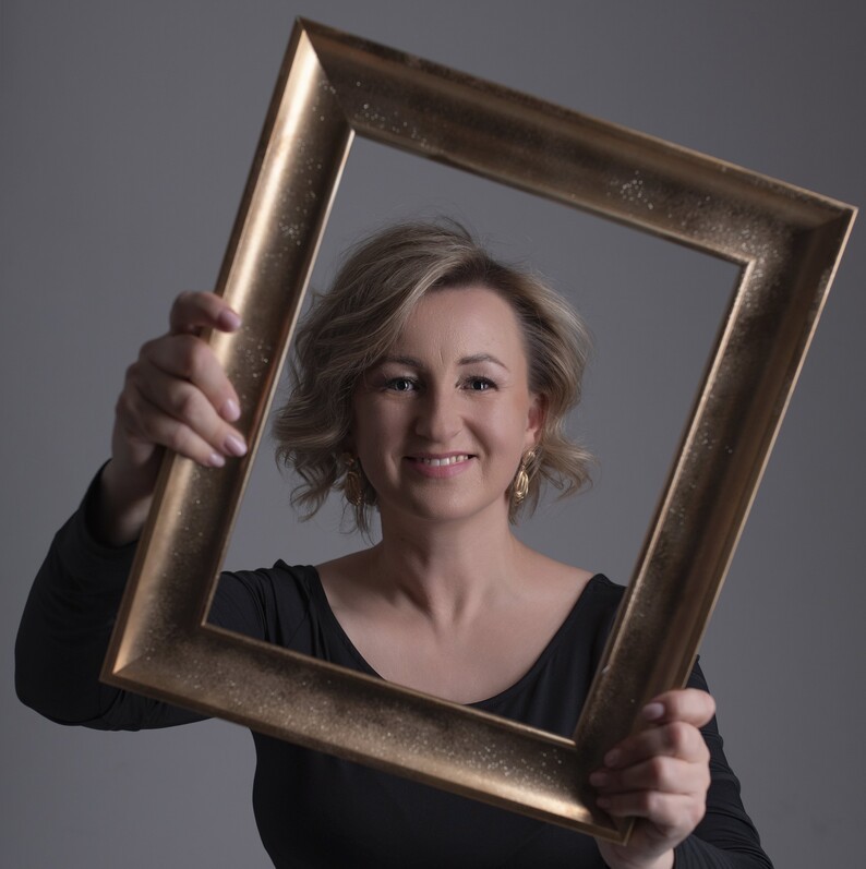 Uśmiechnięta Monika Frys-Żurek trzyma przed sobą pustą ramę obrazu. Jej twarz znajduje się w środku ramy.