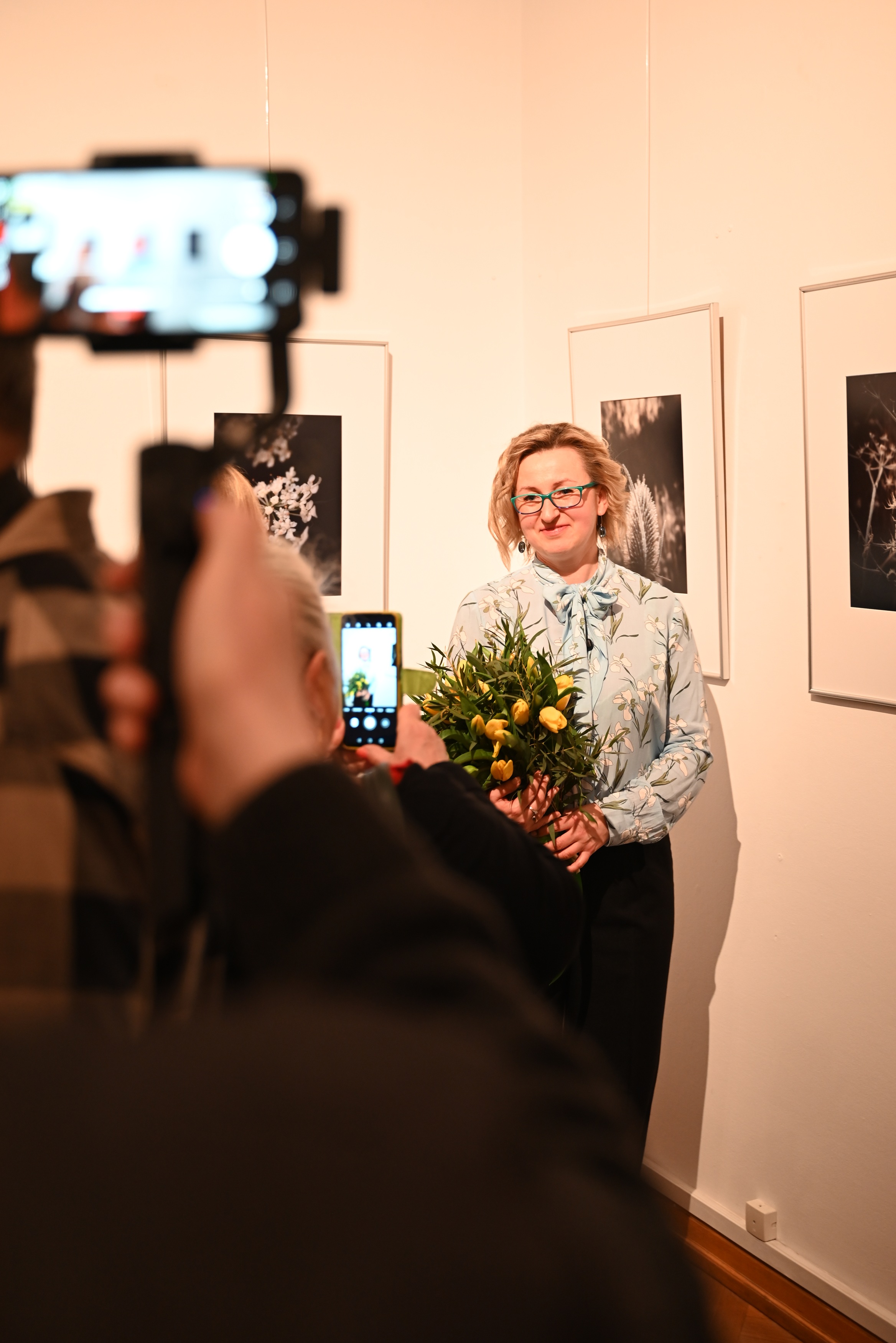 Osoby fotografujące autorkę Monikę Frys-Żurek na tle zdjęć zawieszonych na ścianie.