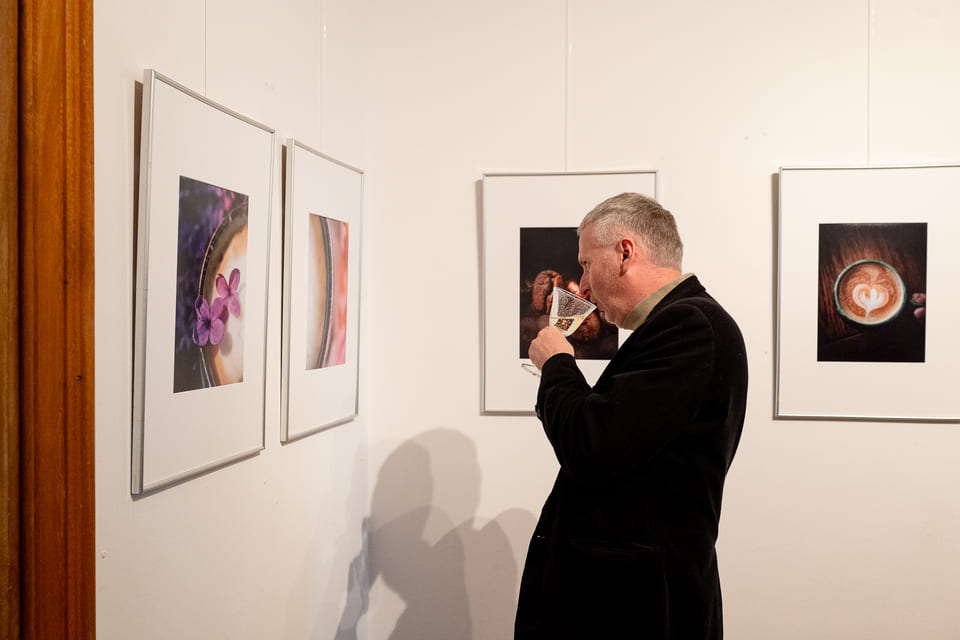 Mężczyzna pijący szampana patrzący na fotografie powieszone na ścianie przed nim.