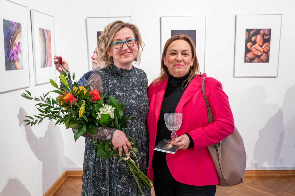 Monika Frys-Żurek trzyma bukiet kwiatów. Obok stoi kobieta. Zdjęcie na tle fotografii prezentowanych na wystawie.