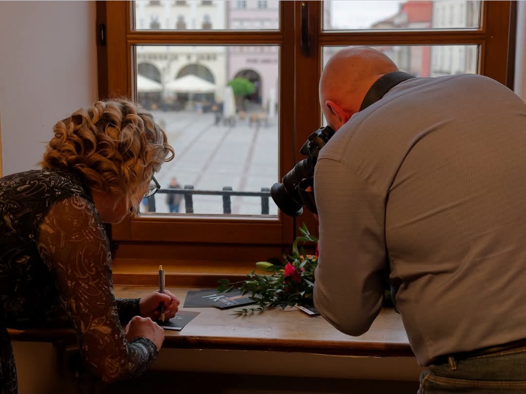 Ujęcie wykonane od tyłu. Monika Frys-Żurek od lewej podpisuje ulotki. Po prawej fotograf robi jej zdjęcie.