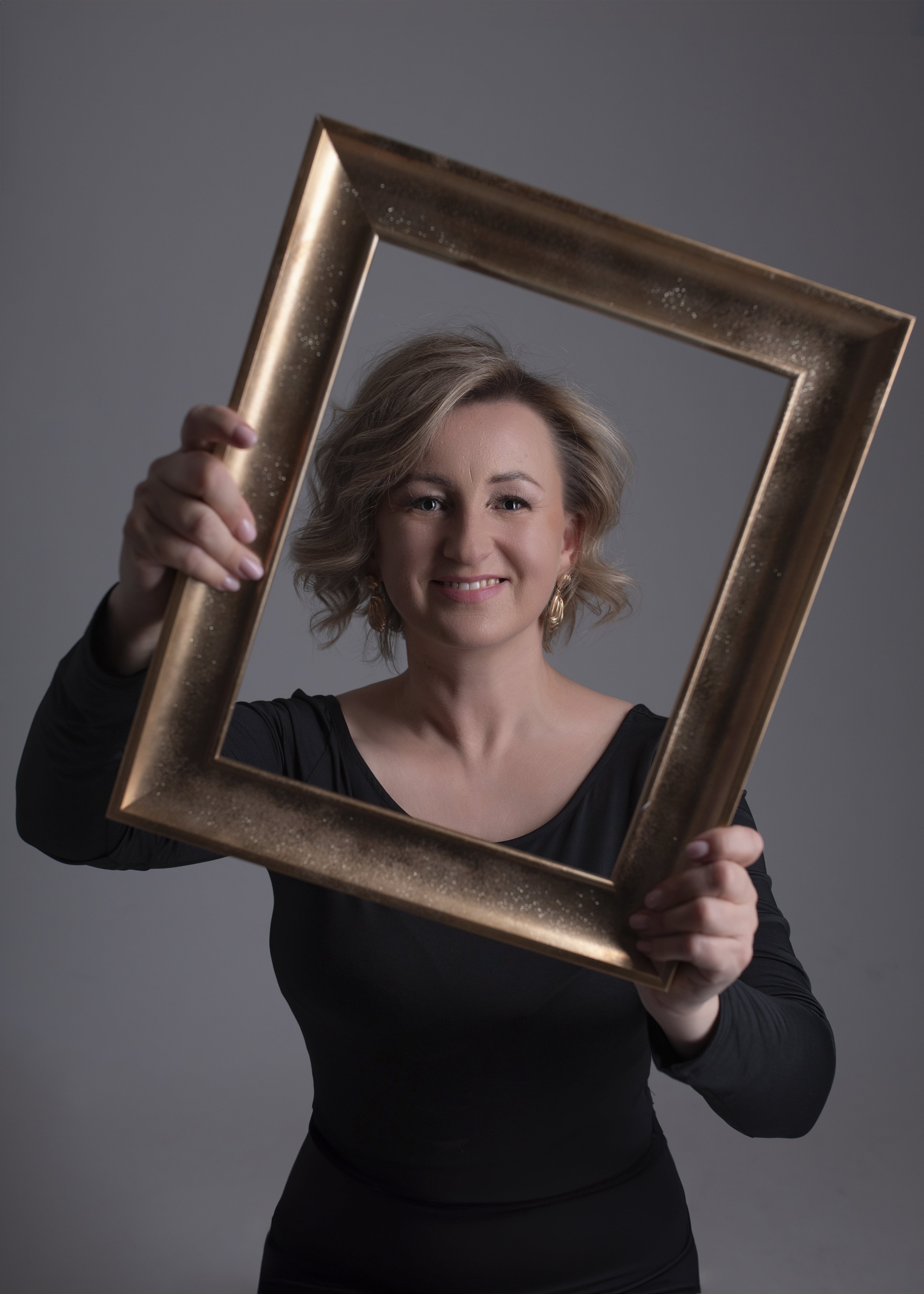 Uśmiechnięta Monika Frys-Żurek trzymająca pustą ramę od obrazu. Jej twarz znajduje się w środku ramy.